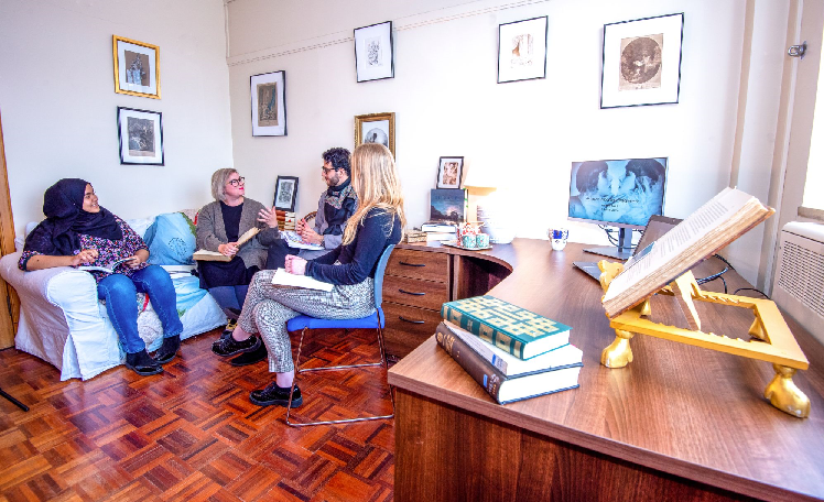 Students sit in an office chatting. In the foreground there are books on the corner of a desk.